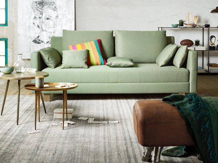JOKA Comoda Doppelliege in Hellgrau mit bunten Zierkissen in einem modernen Wohnbereich mit Couchtisch, Blumen und dekorativen Kunstwerken an der Wand.