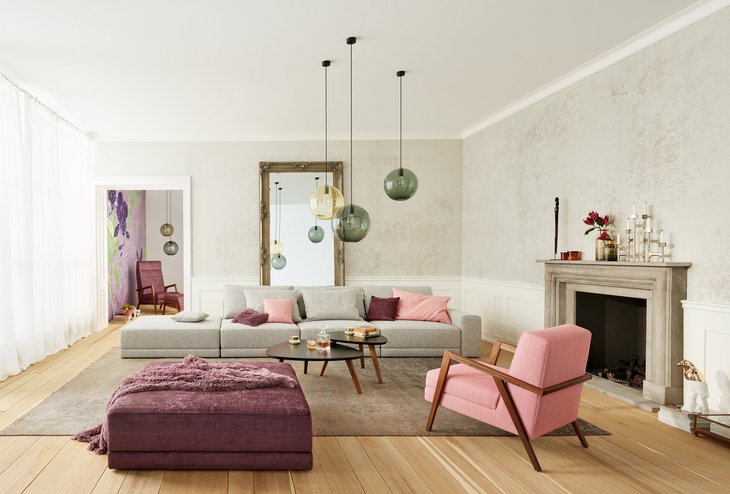 Elegantes, helles Wohnzimmer mit einer großzügigen grauen Wohnlandschaft der Serie Laguna, ergänzt durch rosa Akzente, einen tiefvioletten Hocker und einen Kamin.