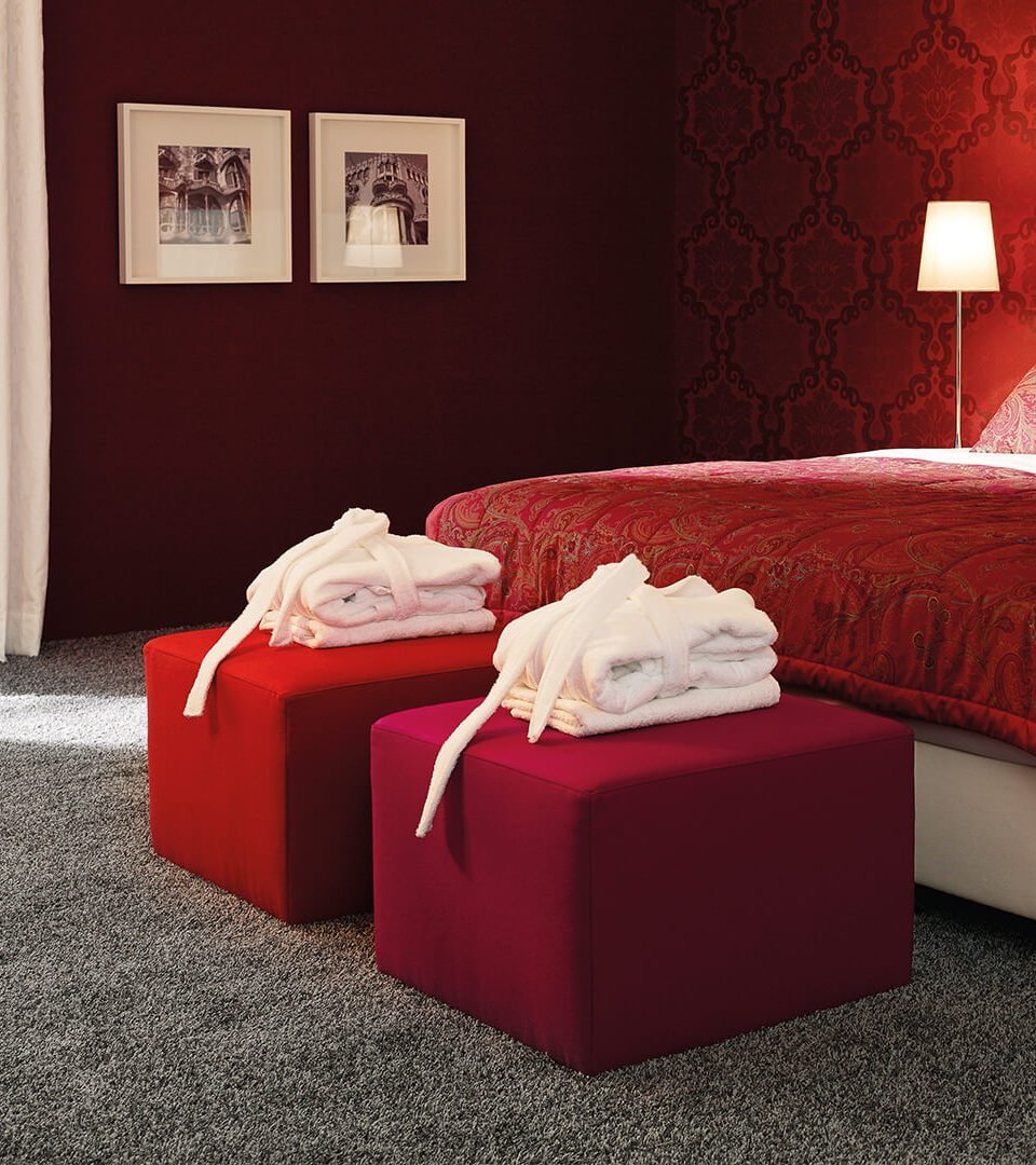 Weißes JOKA Noblesse Boxspringbett mit zwei roten Höckern und roter Wandgestaltung
