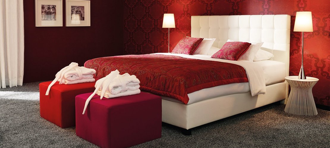 Weißes JOKA Noblesse Boxspringbett mit zwei roten Höckern und roter Wandgestaltung