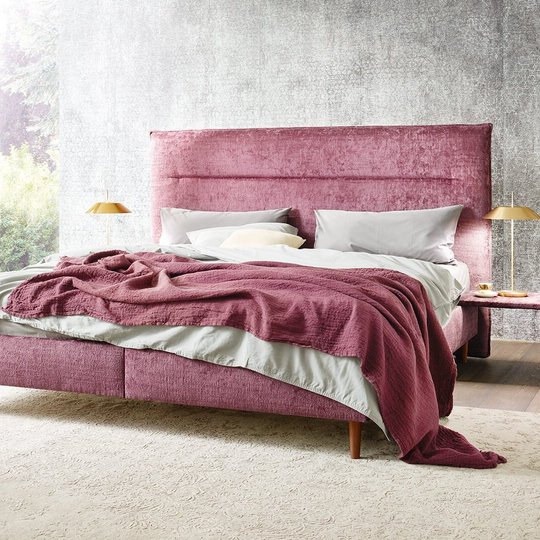 `Gepolstertes JOKA Capri Bett in Beere mit filigranen Holzfüßen, passender Tagesdecke und goldenen Tischlampen vor struktureirter Wand.`