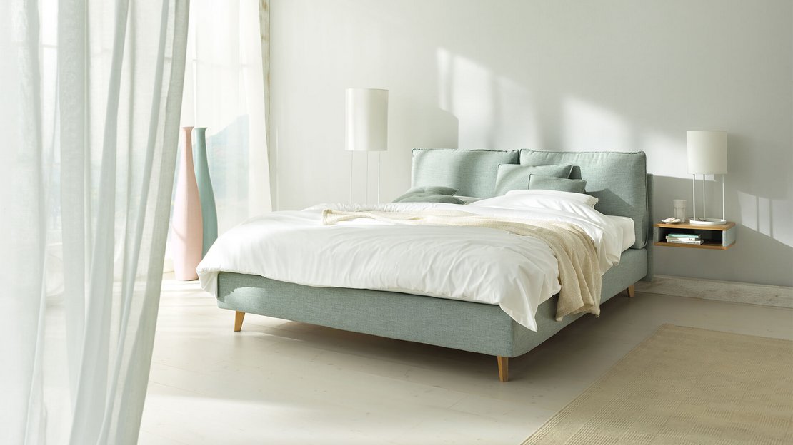 JOKA Arte Boxspringbett in Hellgrün mit weißer Bettwäsche und Holzfüßen im hellen Schlafzimmer