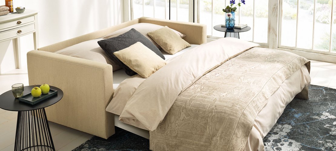 JOKA Magic Längsschläfer umgebaut als komfortables Bett mit beiger Bettwäsche, gemustertem Bezug und dekorativen Kissen in einem hellen Raum mit Teppich und Beistelltisch.