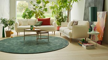 Zwei weiße Bouclé-Sofas (3-Sitzer und 2-Sitzer) über Eck platziert auf grünem Teppich vor einem großen Fenster.