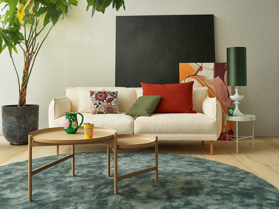 Frontalansicht eines cremeweißen 3-Sitzer-Bouclé-Sofas mit bunten Dekokissen, Beistelltisch und Pflanze.