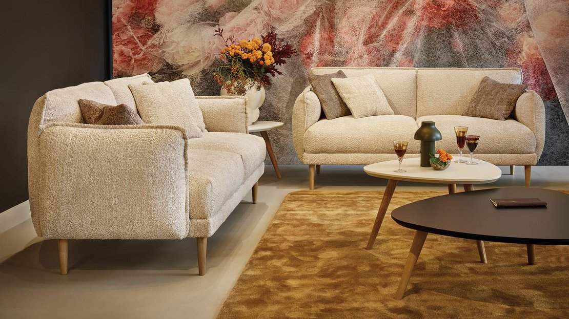 Frontalansicht zweier beigefarbener JOKA Bouclé-Sofas im Loungebereich mit abgestimmten Zierkissen und modernem Beistelltisch-Ensemble.