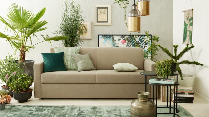 Beigefarbenes JOKA Marea Schlafsofa mit Zierkissen in einem grün bepflanzten Wohnzimmer
