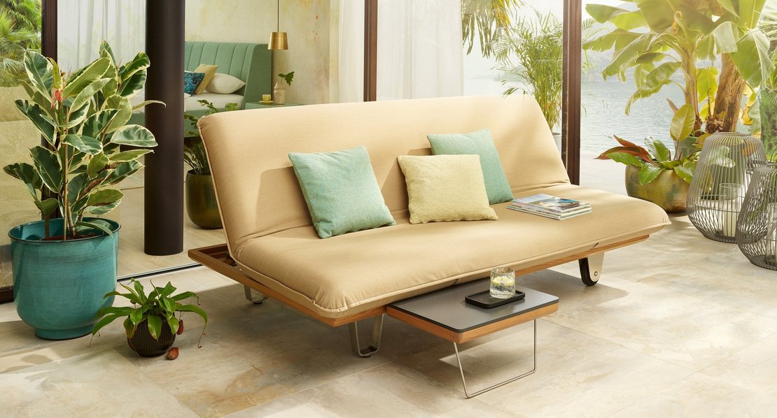 JOKA Butterfly Sunbed in Bankstellung mit einem integrierten kleinen Tisch vor einer Glasfront mit Blick auf Pflanzen und Wasser, dekoriert mit Kissen in Türkis und Hellgrün.