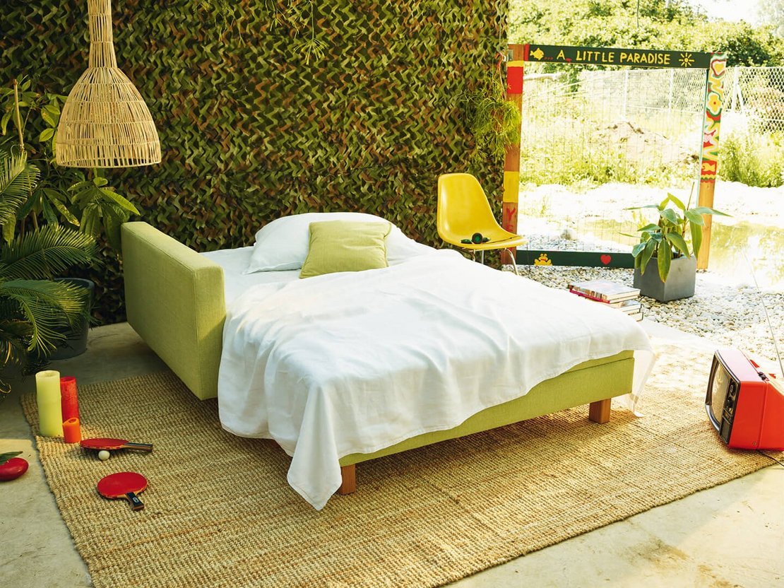 JOKA Quadro Schlafsofa in Hellgrün, ausgeklappt als Bett mit weißer Bettwäsche und Kissen.