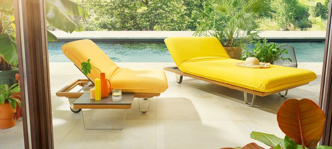 Das Bild zeigt zwei gelbe **JOKA Butterfly Sunbeds** im Freien an einem Pool: eine Liege mit hochgeklapptem Kopfteil und integriertem Tisch auf der linken Seite, und eine zweite, flachere Liege mit einer Sonnenbrille und einem Hut darauf auf der rechten Seite.