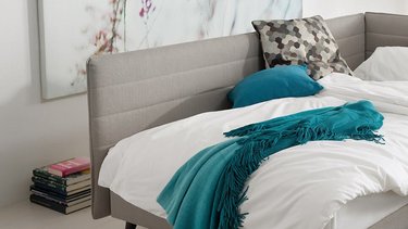 Als Bett hergerichtete grau-beige JOKA Toscana Einzelliege mit weißem Bettzeug und bunten Kissen in einem hellen Wohnraum