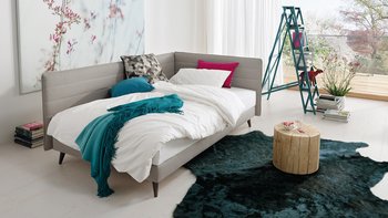 Als Bett hergerichtete grau-beige JOKA Toscana Einzelliege mit weißem Bettzeug und bunten Kissen in einem hellen Wohnraum