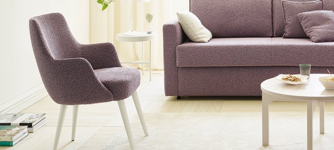 JOKA Paolo Stuhl und Onda Schlafsofa mit violettem Stoffbezug in einem hellen Wohnzimmer mit weißem Couchtisch, hellen Vorhängen und Pflanzen.