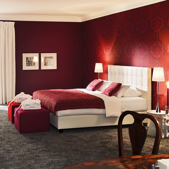 Weißes JOKA Noblesse Boxspringbett in einem eleganten rot gestalltenen Hotelzimmer mit Höcker, Sessel und Sofa