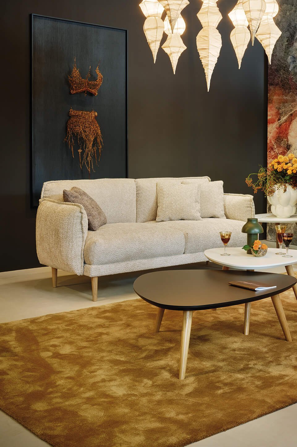 JOKA Loungebereich mit beigefarbenem und senfgelbem Sofa, Bouclé-Sessel und zwei nierenförmigen Beistelltischen auf braunem Teppich vor einer Wand mit Blumenmotiv.