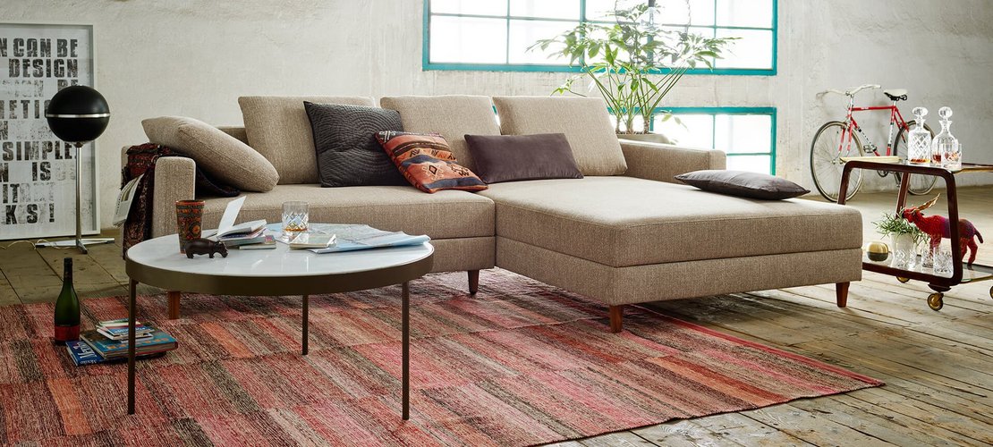 Modernes Loft-Wohnzimmer mit einer beigen Eckcouch inklusive Recamiere, rundem Couchtisch und verputzter Wand im Hintergrund.