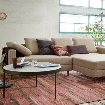 Modernes Loft-Wohnzimmer mit einer beigen Eckcouch inklusive Recamiere, rundem Couchtisch und verputzter Wand im Hintergrund.