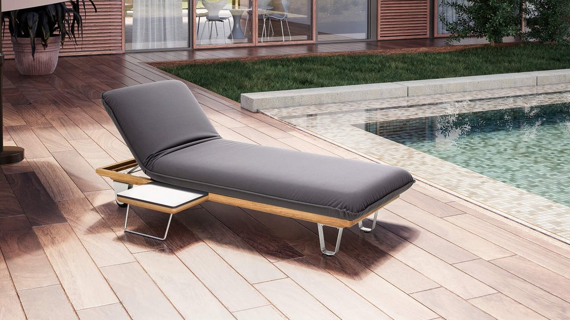 Das Bild zeigt das JOKA Butterfly Sunbed (Einzelliege) in dunklem Grau auf einer Holzterrasse im Freien, platziert neben einem Pool. Das Kopfteil ist in eine Relaxposition hochgeklappt, und an der Seite befindet sich ein kleiner, integrierter Tisch. Im Hintergrund sind ein großer Sonnenschirm, eine moderne Hausfassade mit Glasfronten und grüne Pflanzen zu sehen.