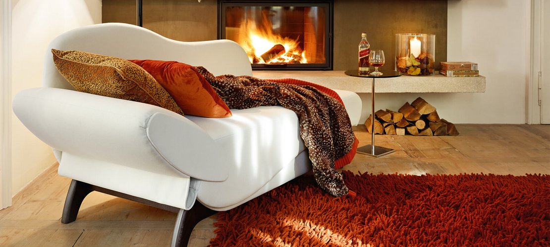 Geschwungenes Recamiere-Sofa in Weiß mit dunklen Holzbeinen und rustikaler Decke vor Kamin