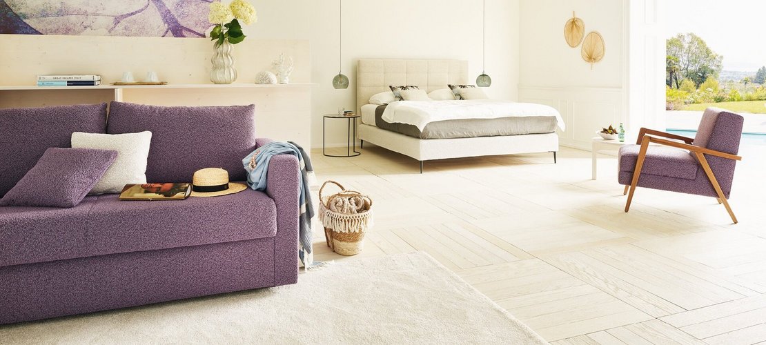 JOKA Mattina Bett in Beige mit lila Schlafsofa Onda und Sesseln in einer lichtdurchfluteten Suite