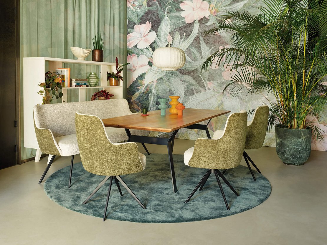 Elegantes JOKA Speiseseparee mit Holztisch, dreifarbigen Vasen, zweifarbiger Polsterbank und Armlehnenstühlen auf rundem blau-grünem Teppich vor einer floralen Fototapete.