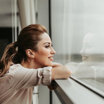 Eine lächelnde Frau stützt sich auf eine Fensterbank und blickt nach draußen durch eine große Fensterscheibe.