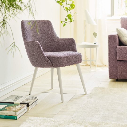 JOKA Paolo Stuhl mit violettem Stoffbezug und weiß lasierten Holzfüßen neben einem passenden Sofa in einem hellen Wohnraum mit Pflanzen.