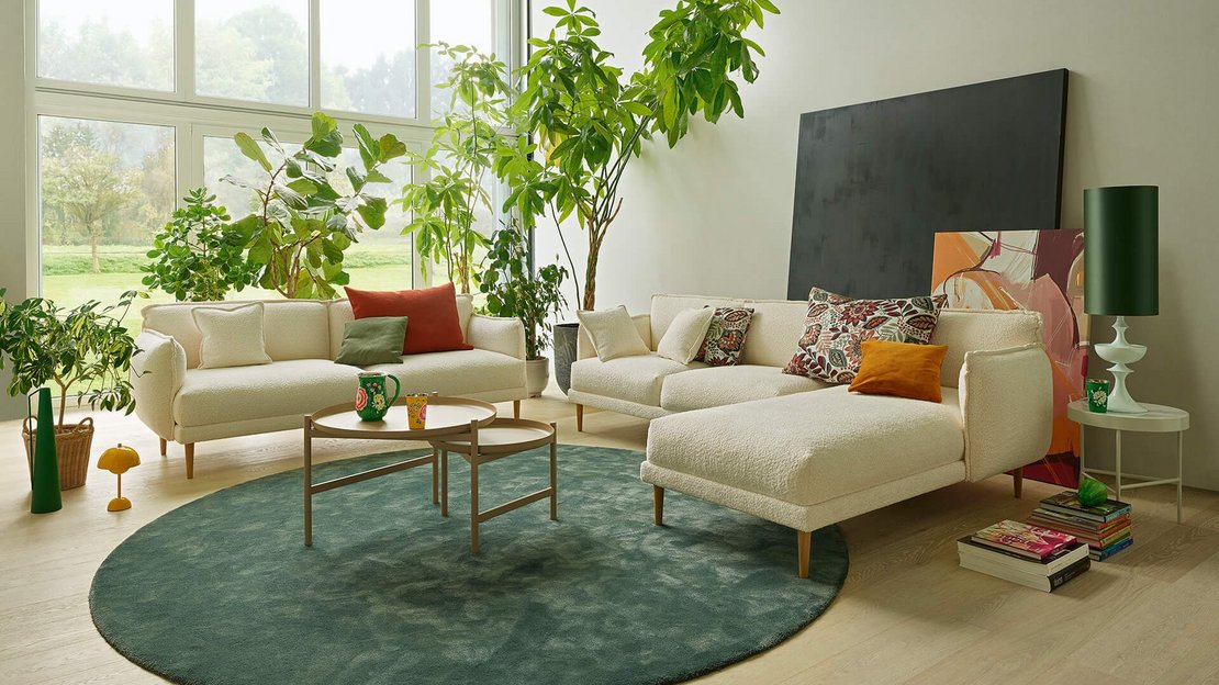 Heller Wohnbereich mit zwei Bouclé-Sofas (3-Sitzer und 2-Sitzer mit Chaiselongue), Couchtisch, grünem Teppich und großen Zimmerpflanzen.