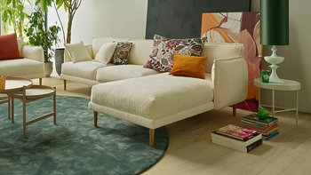 Heller Wohnbereich mit zwei Bouclé-Sofas (3-Sitzer und 2-Sitzer mit Chaiselongue), Couchtisch, grünem Teppich und großen Zimmerpflanzen.