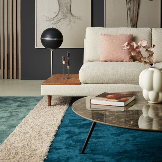 Hochformatige Nahaufnahme eines hellen Sofas mit einer integrierten seitlichen Holzablage, einer Kugel-Stehlampe und Dekoration auf einem blauen Teppich.