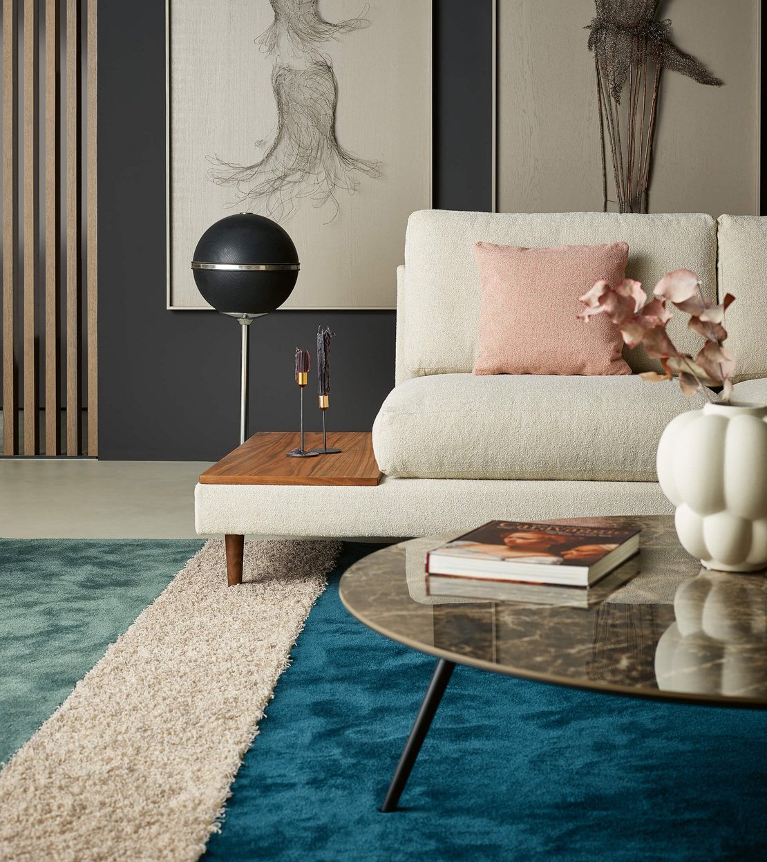 Hochformatige Nahaufnahme eines hellen Sofas mit einer integrierten seitlichen Holzablage, einer Kugel-Stehlampe und Dekoration auf einem blauen Teppich.