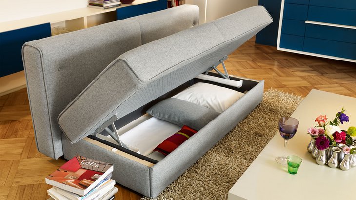JOKA Comoda Doppelliege im vollen Format mit geöffnetem Bettkasten, der praktischen Stauraum für Kissen und Bettwäsche bietet, in einem stilvollen Wohnzimmer.