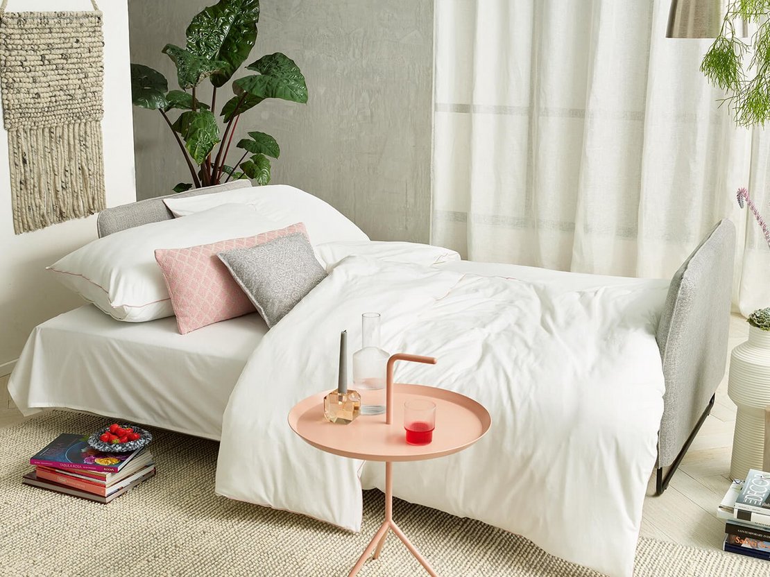 JOKA Lino Doppelliege umgebaut als gemütliches Bett mit weißer Bettwäsche, rosa und grauen Kissen in einem hellen Wohnraum.