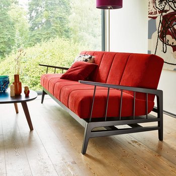 JOKA Cento Kippcouch in Rot mit schwarzem Holzgestell, aufgenommen im Hochformat mit Fokus auf die Armlehne vor einer großen Fensterfront und einem abstrakten Kunstwerk.
