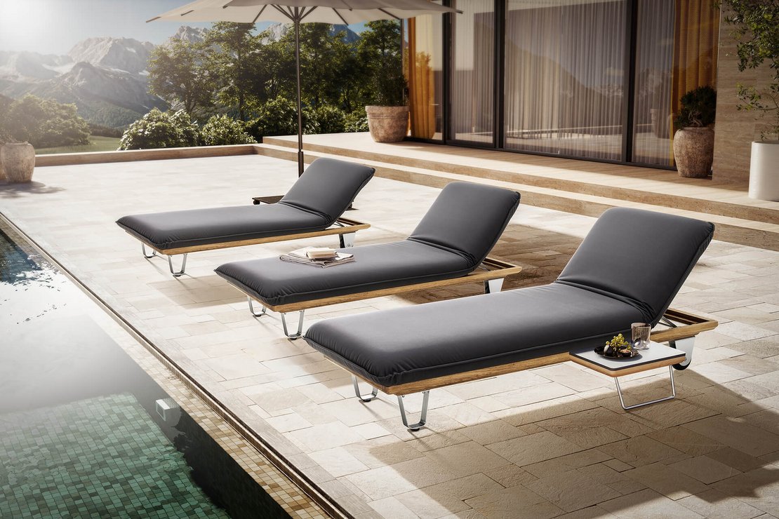 Das Bild zeigt das JOKA Butterfly Sunbed (Einzelliege) mit einer hellgrauen Wetterschutzhülle im Freien auf einer Holzterrasse direkt am Pool, während im Hintergrund eine moderne Architektur mit Glasfronten und Sonnenschirm zu sehen ist.
