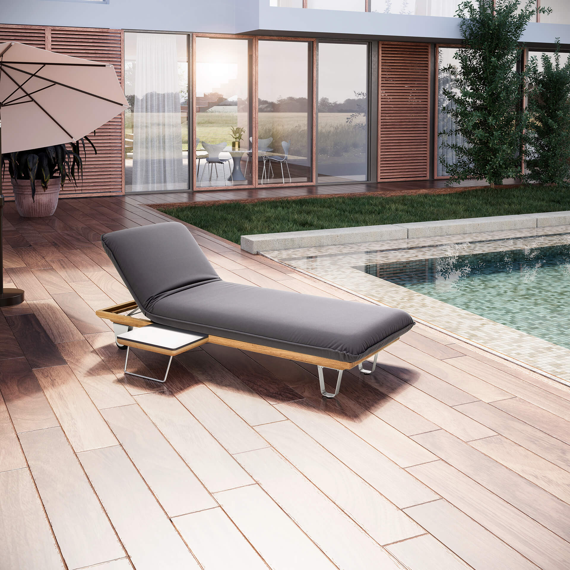 Das Bild zeigt das JOKA Butterfly Sunbed (Einzelliege) in dunklem Grau auf einer Holzterrasse im Freien, platziert neben einem Pool. Das Kopfteil ist in eine Relaxposition hochgeklappt, und an der Seite befindet sich ein kleiner, integrierter Tisch. Im Hintergrund sind ein großer Sonnenschirm, eine moderne Hausfassade mit Glasfronten und grüne Pflanzen zu sehen.